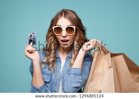 Portrait of a cheery beautiful girl wearing dress and sunglasses holding shopping bags and showing credit card isolated over blue background