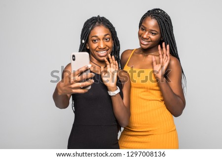 Portrait of two african young girls in dresses make video call on smartphone and wave on camera over white background