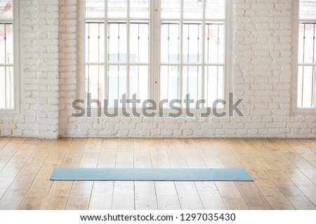Unrolled yoga mat lying in empty light fitness studio on wooden floor, unfolded sport equipment prepared for training, modern pilates loft room ready for workout session. Healthy life concept
