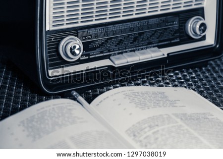 Vintage radio of the 60s on a tablecloth with metallic weft, front view, dark background, open book and pencil resting on top, monochrome interpretation of the shot.