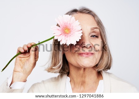 Cheerful elegant elderly woman holding a flower near the face on a gray background Royalty-Free Stock Photo #1297041886