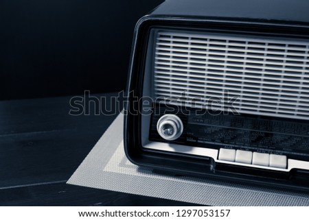 Vintage radio from the 60s on the Wengè wooden table with a classic light-colored tablecloth, partial view, free area on the left side with black background, monochrome interpretation of the shot. Royalty-Free Stock Photo #1297053157