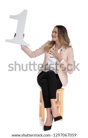 happy businesswoman sits on wooden chair on white background and looks to side at number one sign, full body picture