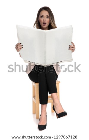 shocked businesswoman sits on wooden stool while holding newspaper on white background, full length picture