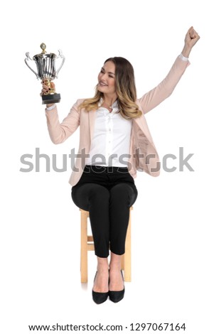 happy businesswoman looks at trophy to side and celebrates with hand in the air while sitting on wooden cahir on white background, full length picture