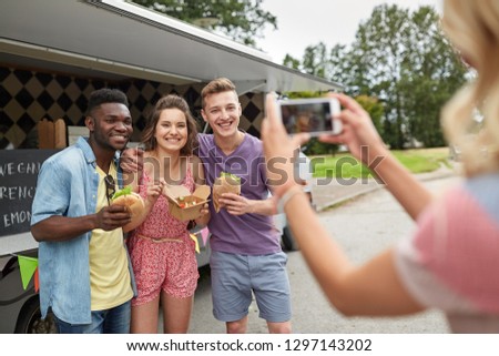 leisure, technology and people concept - young woman taking picture of her happy friends eating hamburgers and wok at food truck