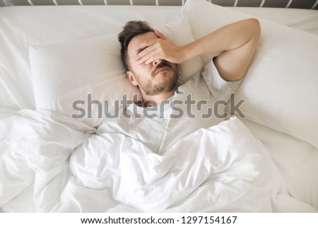 Tired young man in bed closing his eyes before waking up. Hangover morning. Sleep disorder and problems.  Royalty-Free Stock Photo #1297154167