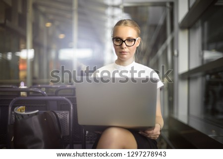 Successful woman entrepreneur in glasses checking e-mail on portable laptop computer while sitting in airport terminal before important business trip abroad. Female in spectacles using netbook 