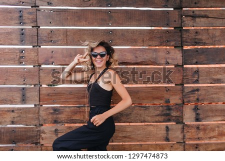Inspired tanned woman expressing energy while posing outdoor. Photo of laughing young lady in black clothes isolated on wooden background.