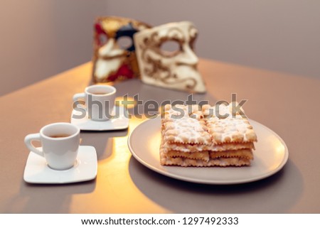 Typical Italian carnival fritters (Chiacchiere di Carnevale) dusted with powdered. Composition with two cups of coffee and in the background are Venetian carnival masks.