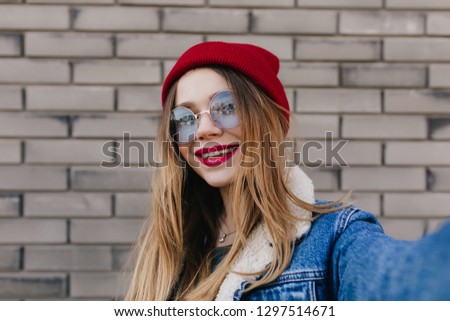 Interested woman with light-brown hair making selfie and smiling on the street. Outdoor photo of pretty caucasian female model in denim jacket taking picture of herself.
