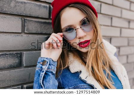 Close-up shot of magnificent woman with light-brown hair touching her blue glasses. Outdoor photo of charming girl in denim jacket standing on urban background in cold morning.