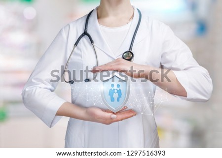 A young doctor is holding a big shield with group of people or family members icon inside between her palms in protective gesture. The family medicine and life and health insurance concept.