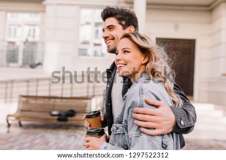 Interested girl drinking coffee during walk with boyfriend. Outdoor photo of happy young woman enjoying date.