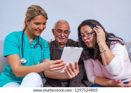 Positive senior couple with their caregiver in nursing home for elderly. Elder married couple sitting on the couch and smiling at nurse with a tablet computer