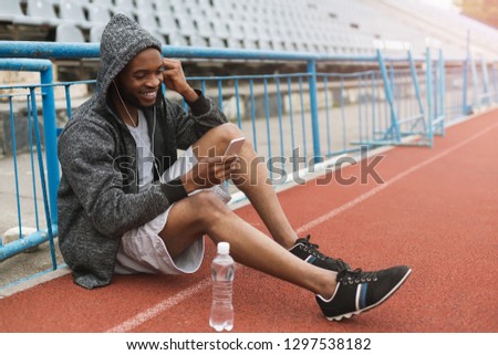 Runner black man listening to music with earphones on stadium, having respite.