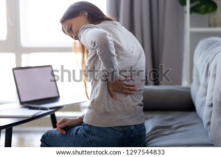 Young woman feeling pain in spine back after sedentary computer work sitting in bad posture on sofa at home, tired girl rubbing backache tensed muscles suffers from lower lumbar kidney ache, backpain Royalty-Free Stock Photo #1297544833