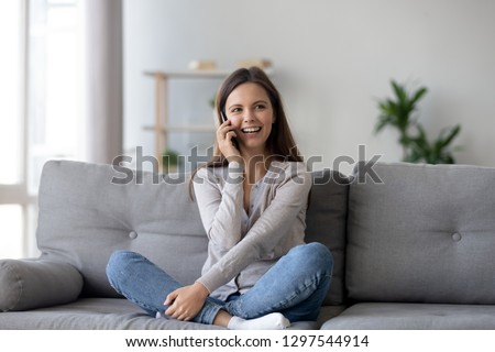 Happy young woman caller talking on the phone at home, cheerful teen girl enjoys pleasant mobile conversation, smiling millennial female holding cell speaking making call by telephone in living room Royalty-Free Stock Photo #1297544914