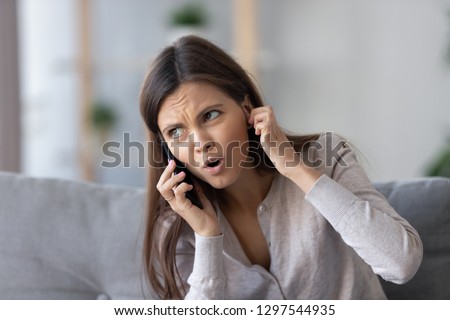 Annoyed young woman talking on the phone not hearing bad signal lost no mobile connection on cellphone, irritated girl closing ear frustrated by communication telecommunication service problem indoor Royalty-Free Stock Photo #1297544935