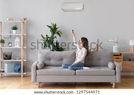Young happy woman switching on air conditioner sitting on couch at convenient cozy home, lady relaxing on sofa in living room holding remote climate control to cooler system set comfort temperature Royalty-Free Stock Photo #1297544971