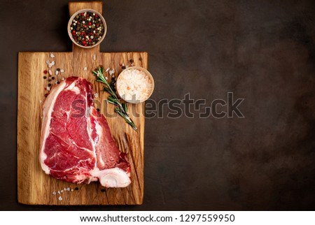 
Raw meat steak with seasoning on a cutting board, background concrete. Space for text. Beef T-bone steak, top view. Barbecue concept. Ingredients for roasting meat.