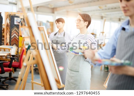 Side view portrait of inspired young woman painting picture on easel enjoying work with group of students in art studio