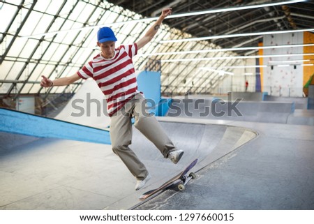 Skilled young parkour boy in t-shirt, pants and baseball cap practicing jump on skateboard over descent at stadium