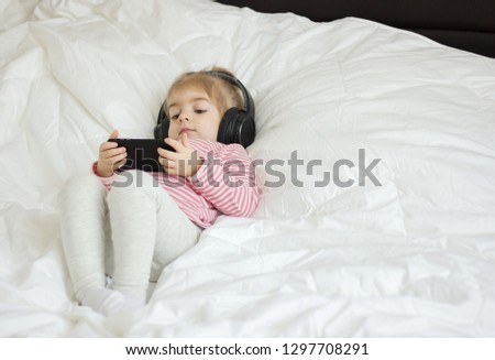 Cute little girl lying on the bed and listening to music using smartphone and listening on earphones .
