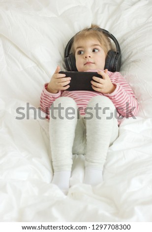 Cute little girl lying on the bed and listening to music using smartphone and listening on earphones .