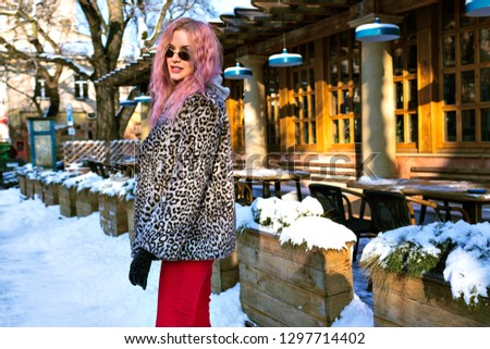 Winter outdoor fashion portrait of stylish young woman posing at snowy European city, grunge London street wear, unusual pink hairs, trendy leopard jacket, vintage glasses, rock vibes.