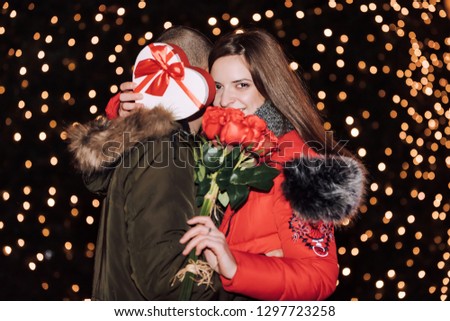 Couple. Love. Holiday. Outdoors. Woman is holding a gift box and bouquet of roses and smiling while hugging her man at night in the city street - Image