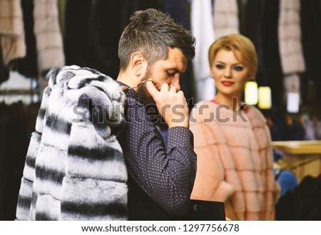 Woman chooses expensive sable overcoat. Shop assistant with beard and woman select furry coat. Finance and shopping concept. Man and girl with calm faces hold furry coats on clothes rack background.