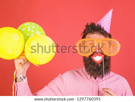 Hipster in giant sunglasses celebrating. Guy in party hat with air balloons celebrates. Photo booth fun concept. Man with beard on cheerful face holds smiling mouth on stick, red background.