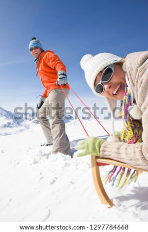 Couple on winter vacation with man pulling wife through snow on sled smiling at camera