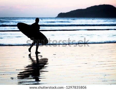 Unidentified surfer walking on the seashore after a day surfing photographed in black and white.