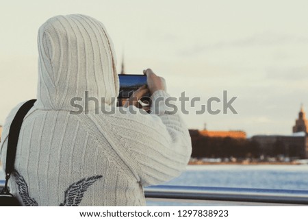 Man in white knitted jacket with hood taking photo of cityscape with his smartphone in Riga, Latvia