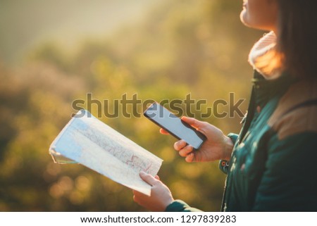 Crop picture of a girl holding a map and mobile phone trying to plan the route in the mountains while hiking and walking in the forest, flare warm sunset light