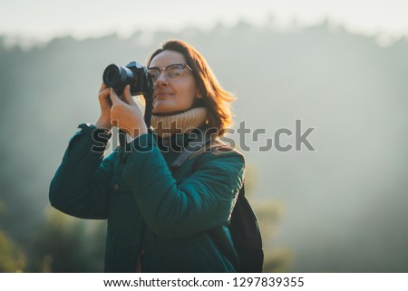 Hipster brunette girl making pictures of the mountains on her modern professional photo camera while travelling in the forest, sun and blurred nature on the background