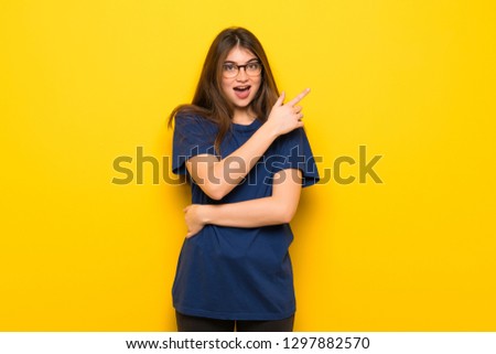 Young woman with glasses over yellow wall surprised and pointing side