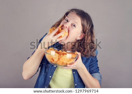 Portrait of a cute young girl with a glass bowl full of chips puts a lot of chips in her mouth.