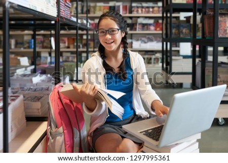 Smiling schoolgirl reading book and working on laptop in library