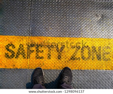 A man standing on “SAFETY ZONE” word.He is wearing jeans with blue Shout. He is standing on Metal floor.  Photo safe and  Comfort Zone concept idea.