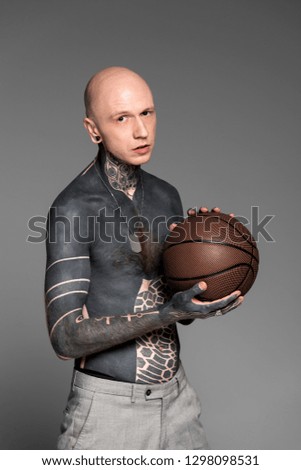 bare-chested man with tattoos holding basketball ball and looking at camera isolated on grey