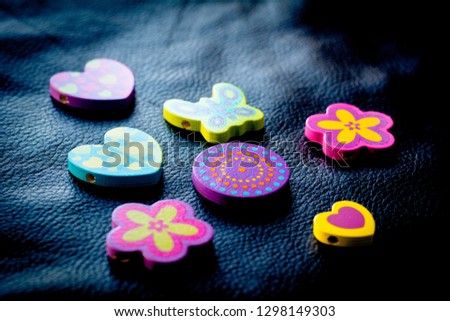Close up of colorful wooden beads in the shape of heart, butterfly, circle and flower, displayed over a black leather background, illuminated by natural light.
