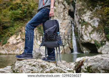 Unidentified man tourist in casual clothes is holding an equipped backpack for travel against the backdrop of a mountain river and stones. Concept of active recreation and comfortable clothing