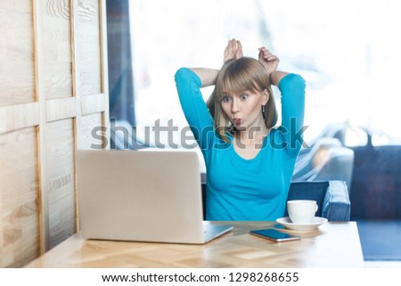 Happy childish young girl freelancer with blonde hair in blue t-shirt are sitting in cafe and making video call on laptop, talking and teased with her friend, holding hands near head like bunny ears.