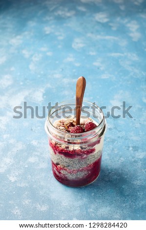 Raspberry chia seed yogurt pudding with muesli and fresh berries. Healthy dessert in glass jar on blue background. Close up view
