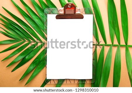 Clipboard with blank piece of paper on big green leaf of parlor palm tree, golden orange gradient table background. Empty sheet on branch of tropical plant. Top view, close up, copy space, flat lay.