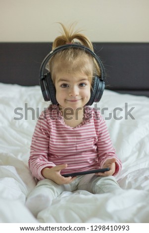 Little girl sitting on the bed , listening music with headphones and use smartphone.