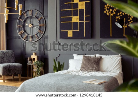 Real photo of dark bedroom interior with double bed with white sheets, grey blanket and open book, paintings on wall with molding and gold lamp Royalty-Free Stock Photo #1298418145
